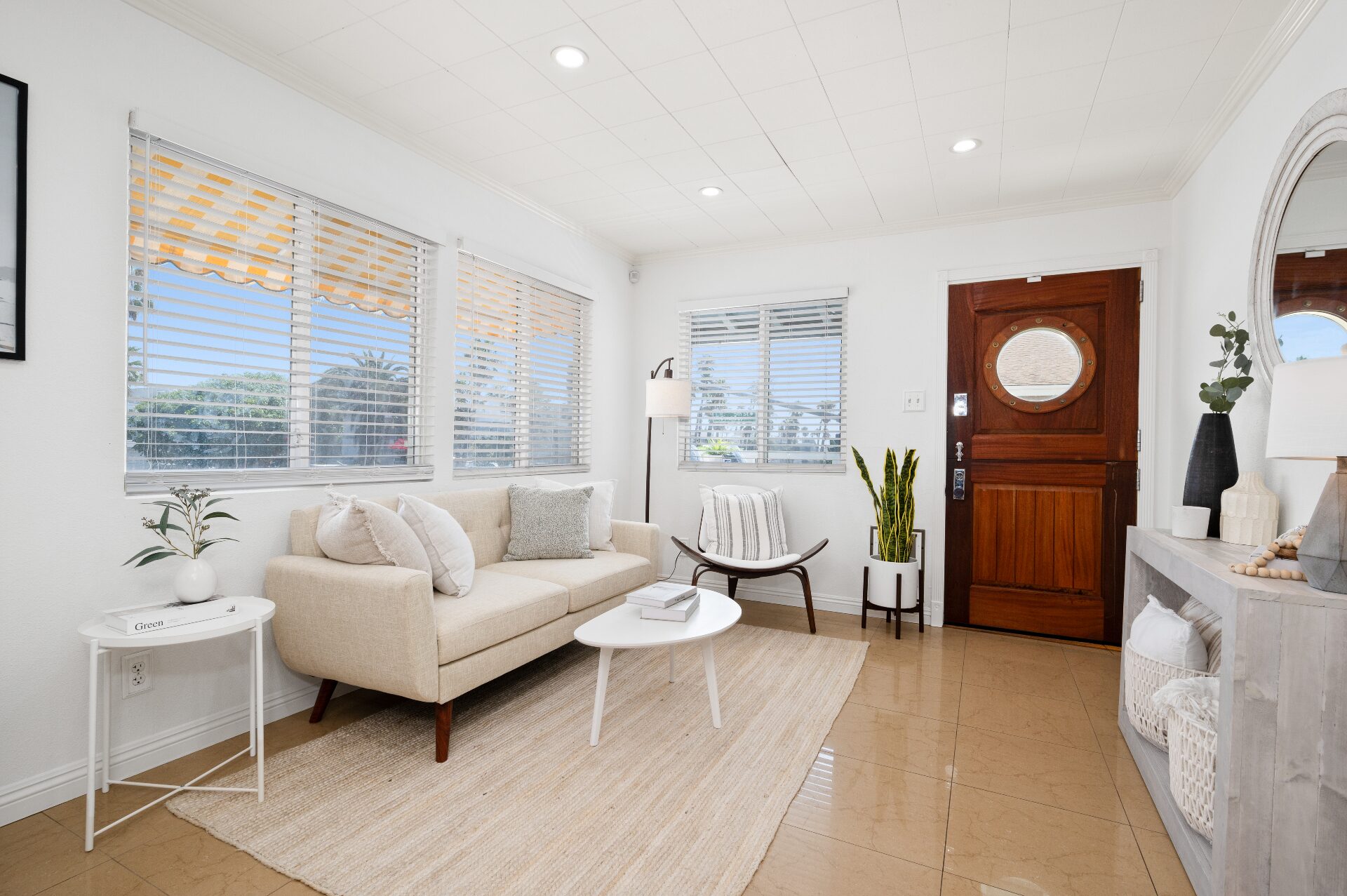 Bright sitting room with porthole wood door, soft neutral seating, woven textures, and palm-lined windows