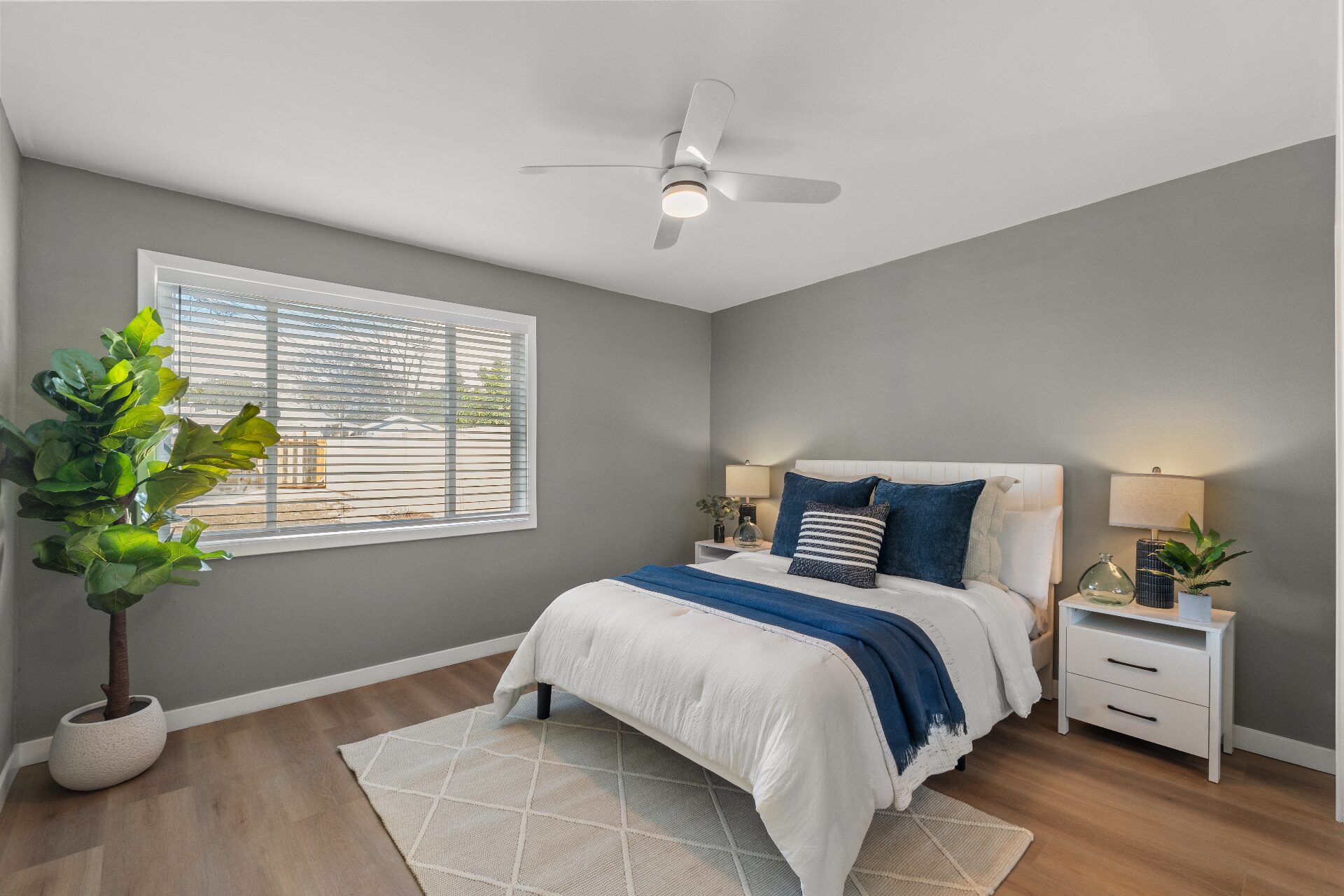 Staged bedroom with neutral walls, navy accents, white linens, and large window with natural light