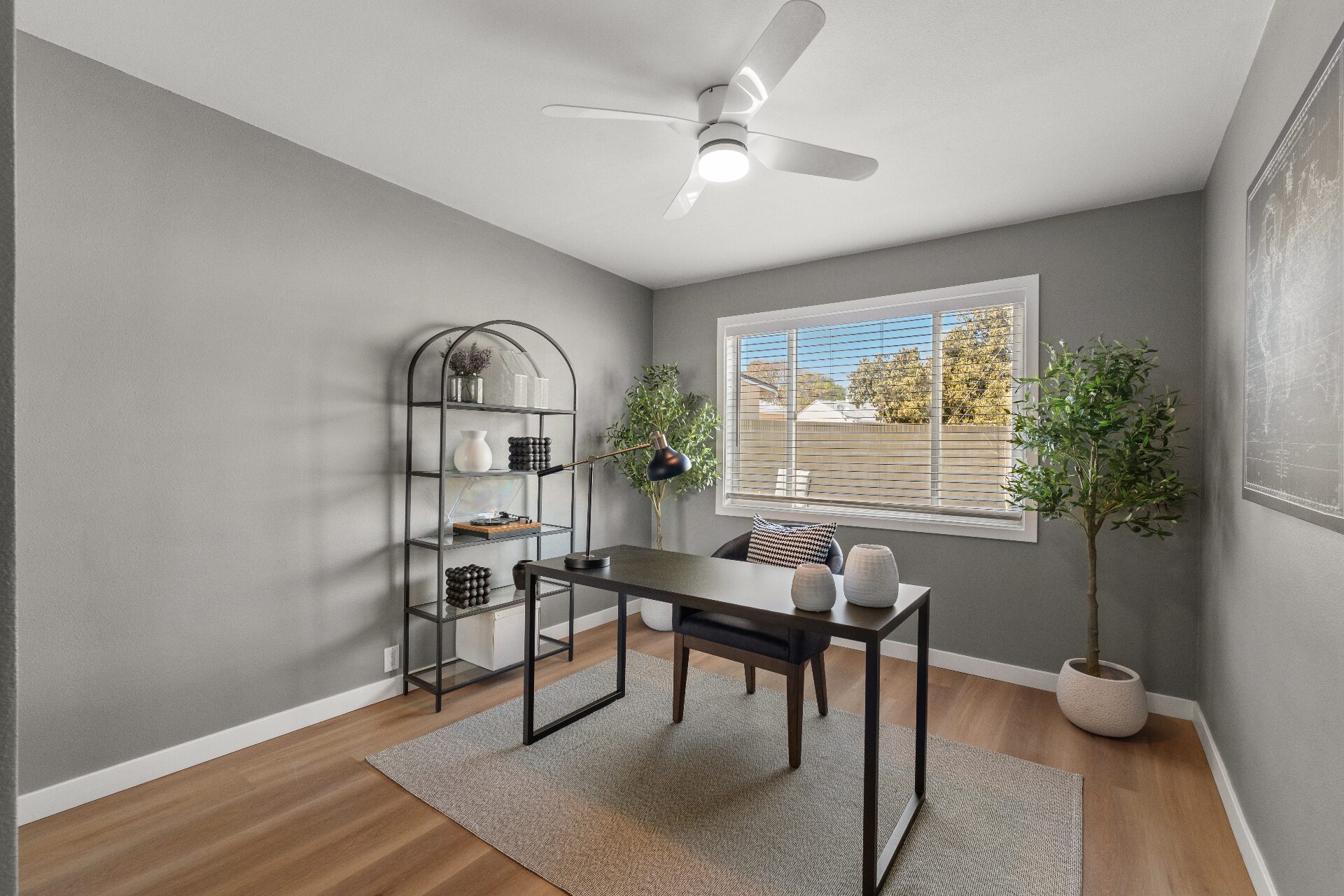 Staged home office with dark wood desk, arched bookshelf, olive trees, and natural light