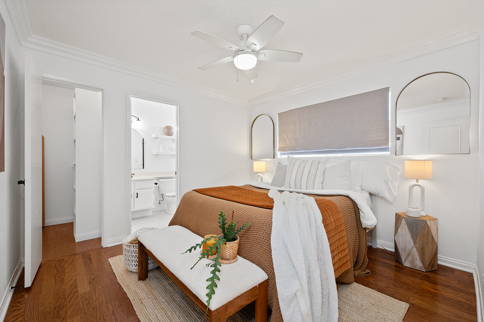 Cozy bedroom with rust-colored throw, layered linens, arched mirrors, and en-suite bath