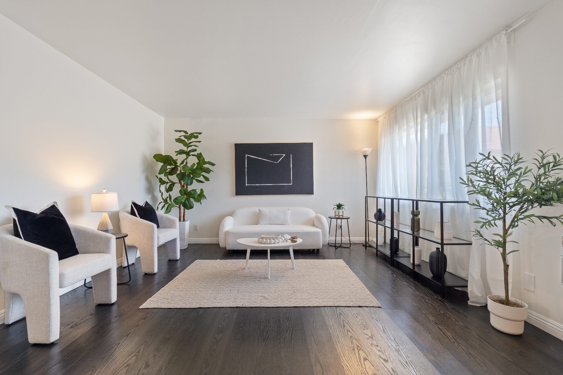 Minimal staged living room with sculptural white seating, black oak floors, soft drapery, and monochrome art