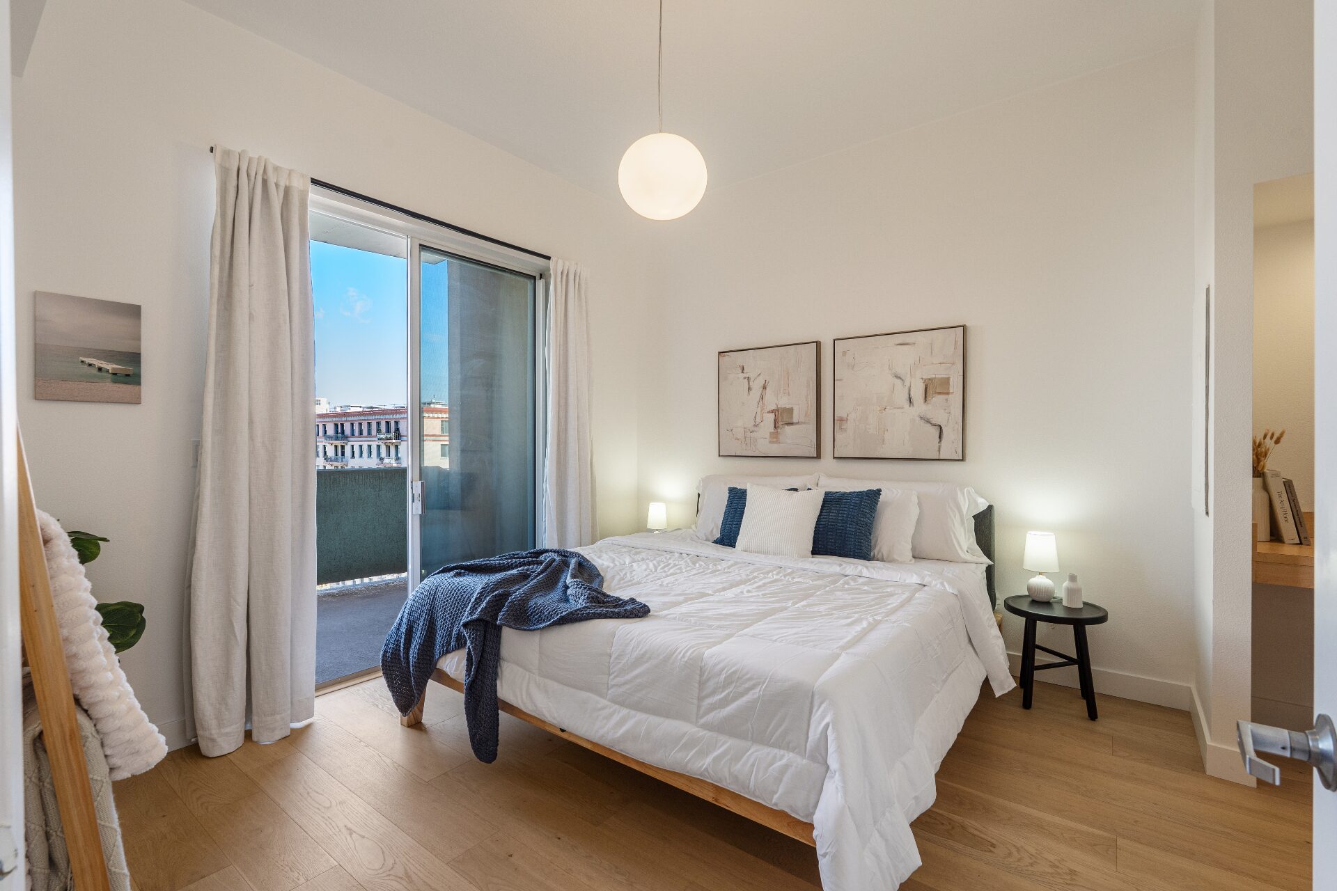 Light-filled bedroom with king bed, abstract art, balcony with city views, and pendant lighting