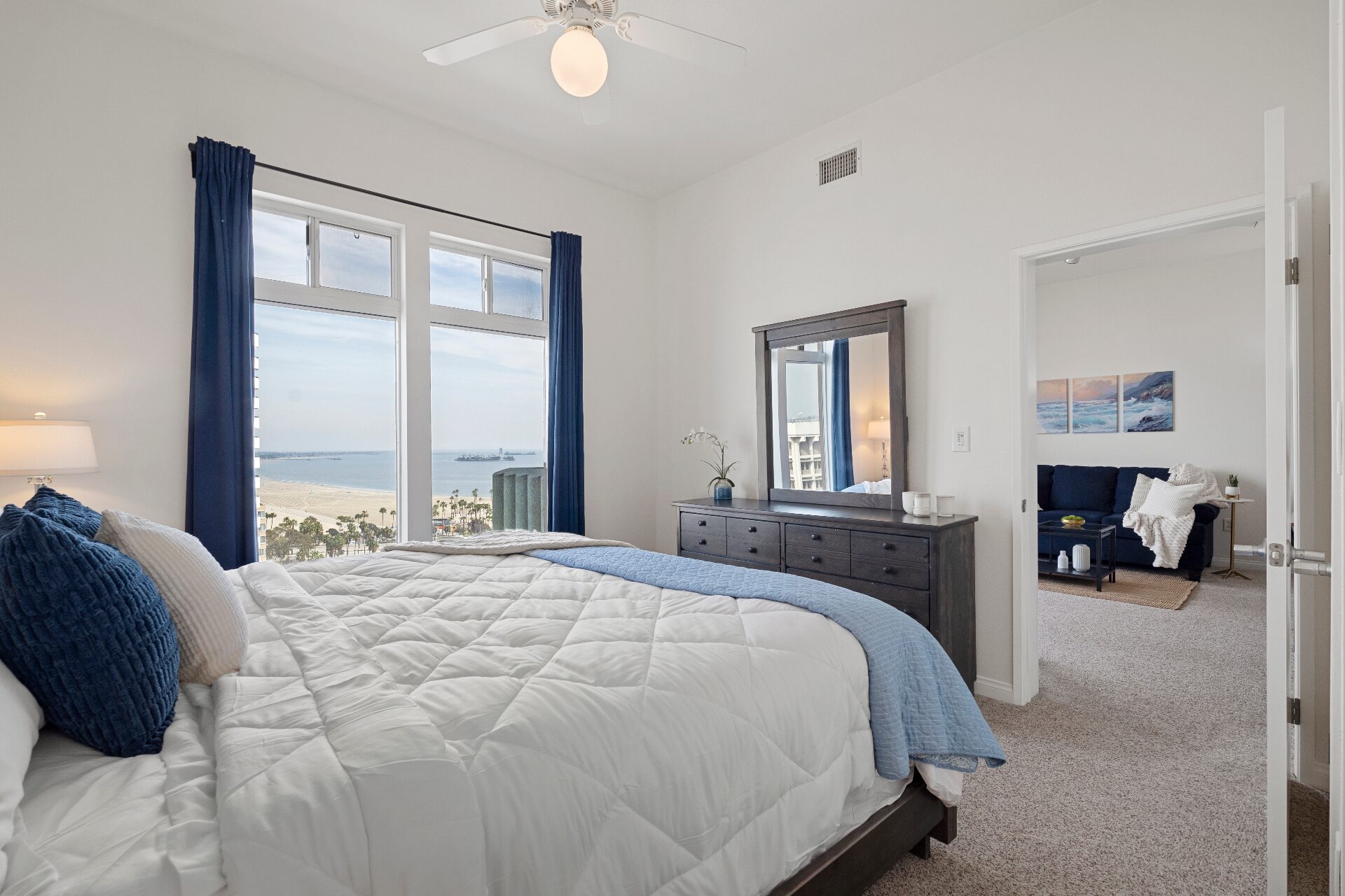 Oceanfront bedroom with floor-to-ceiling windows, navy drapes, and panoramic beach and harbor views